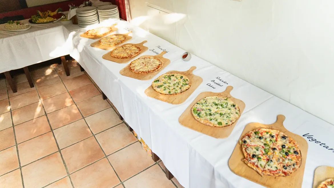 Lange Tafel mit sieben Pizzen auf Holzbrettern, daneben handgeschriebene Sortenschilder